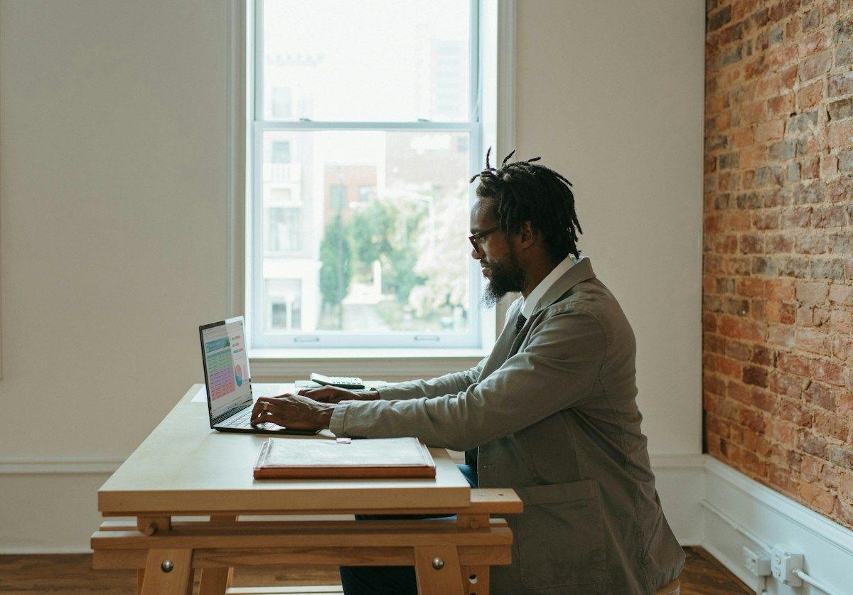 Man working on a laptop at a desk by a bright window